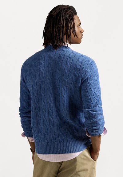 Μπλε πλεκτό πουλόβερ με ραμμένο τελείωμα και μανσέτες, με υφή σχεδίου. Φορεμένο επάνω από πουκάμισο με ροζ ρίγες.