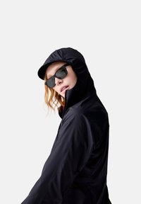 Veste légère noire avec capuche, dotée d'une fermeture éclair sur le devant et d'une texture lisse, associée à des lunettes de soleil noires. Le mannequin pose de côté.