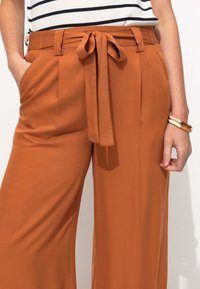 Pantalons larges en tissu orange rouille avec une texture lisse, dotés d'un détail de taille nouée et de poches latérales. Haut rayé visible au-dessus.