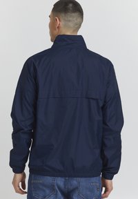Marinblå vindjacka med hög krage, elastiska ärmslut och rak fåll. Slät, lättviktig tyg, minimalistisk design, inga synliga logotyper.