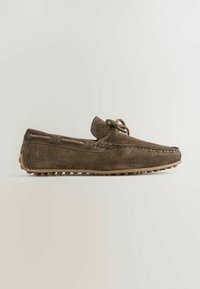 Mocassin en daim marron avec semelle en caoutchouc et lacets noués en nœud sur le dessus, présenté sur un fond neutre.