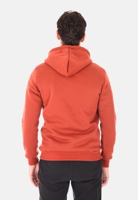 Uomo che indossa una felpa color ruggine e pantaloni neri, in piedi con le spalle alla fotocamera contro uno sfondo bianco semplice.