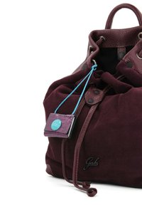 Zaino in camoscio color mogano con dettagli in pelle, chiusura a coulisse e una piccola etichetta viola con un bottone blu, cuciture dettagliate e texture liscia.