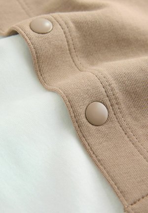 Brunt stof med tekstureret overflade, der har en glat knapdetalje, foldet over et lyst cremefarvet materiale, hvilket skaber et lagdelt udseende.