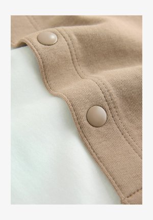 Pellavanvärinen kangas, jossa on teksturoitu pinta ja sileä nappikoriste, taitettuna vaalean kermaiselle kankaalle, luoden kerroksellisen ilmeen.