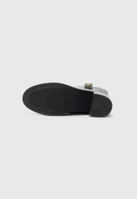 Semelle de chaussure noire tournée vers le haut avec une texture antidérapante et un petit logo doré sur la partie supérieure en cuir gris, le tout sur fond blanc.