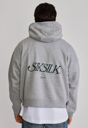 Mężczyzna ubrany w szarą bluzę z kapturem z logo "SIKSILK" na plecach, nałożoną na białą koszulę i niebieskie dżinsy, widziany od tyłu.