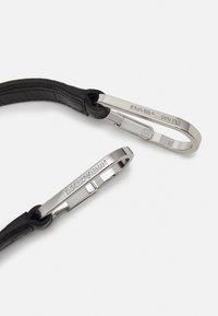 Två silverfärgade metalklips med ingraverad "EMPORIO ARMANI" fästa vid svarta läderremmar mot en vit bakgrund.