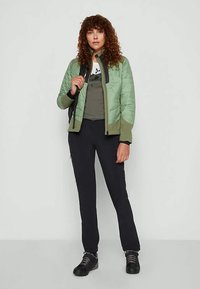 Naine lokkis juustega, seljas roheline tikandiga jakk, oliiviroheline särk, mustad püksid, mustad matkenaistel ja hoiab seljakotirihmast.