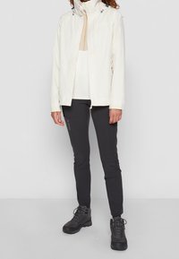 Veste blanche imperméable avec capuche ajustable, associée à un pantalon noir ajusté et des chaussures de randonnée noires. Texture lisse et design minimaliste.