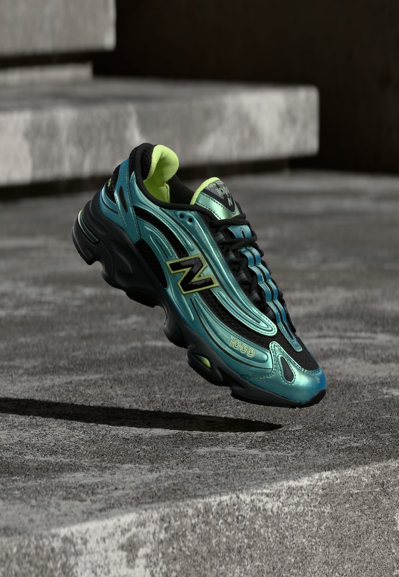 Πράσινο-μπλε και μαύρο αθλητικό παπούτσι New Balance με κίτρινες λεπτομέρειες, που αιωρείται πάνω από μια τσιμεντένια σκάλα σε εξωτερικό χώρο.