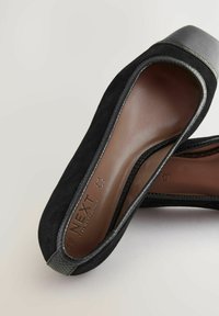 Scarpe slip-on nere in suede con un morbido accento in pelle scura sulla punta e bordi texture. L'interno è di un colore contrastante marrone chiaro.