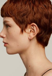 Pendiente de aro de oro con un pequeño diamante; cabello corto, ondulado y marrón rojizo; piel suave con sutilejas pecas, vista de perfil.