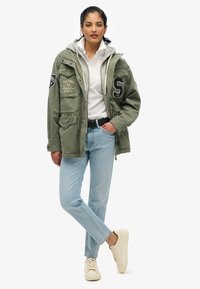 Oliwkowa kurtka w militarnym stylu z kapturem, z naszytymi łatami, w połączeniu z białą koszulą z kołnierzykiem i jasnoniebieskimi dżinsami, kremowymi trampkami.