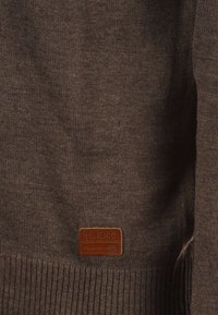 Brun strukturert genser med ribbestrikket kant og en lærslabel med teksten "BLEND GLOBAL JEANMAKER." Bløtt strikkemateriale.