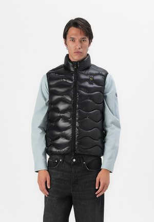 Gilet invernale nero con collo alto, tessuto trapuntato e zip frontale. Indossato sopra una maglietta azzurra chiara, abbinato a jeans scuri.