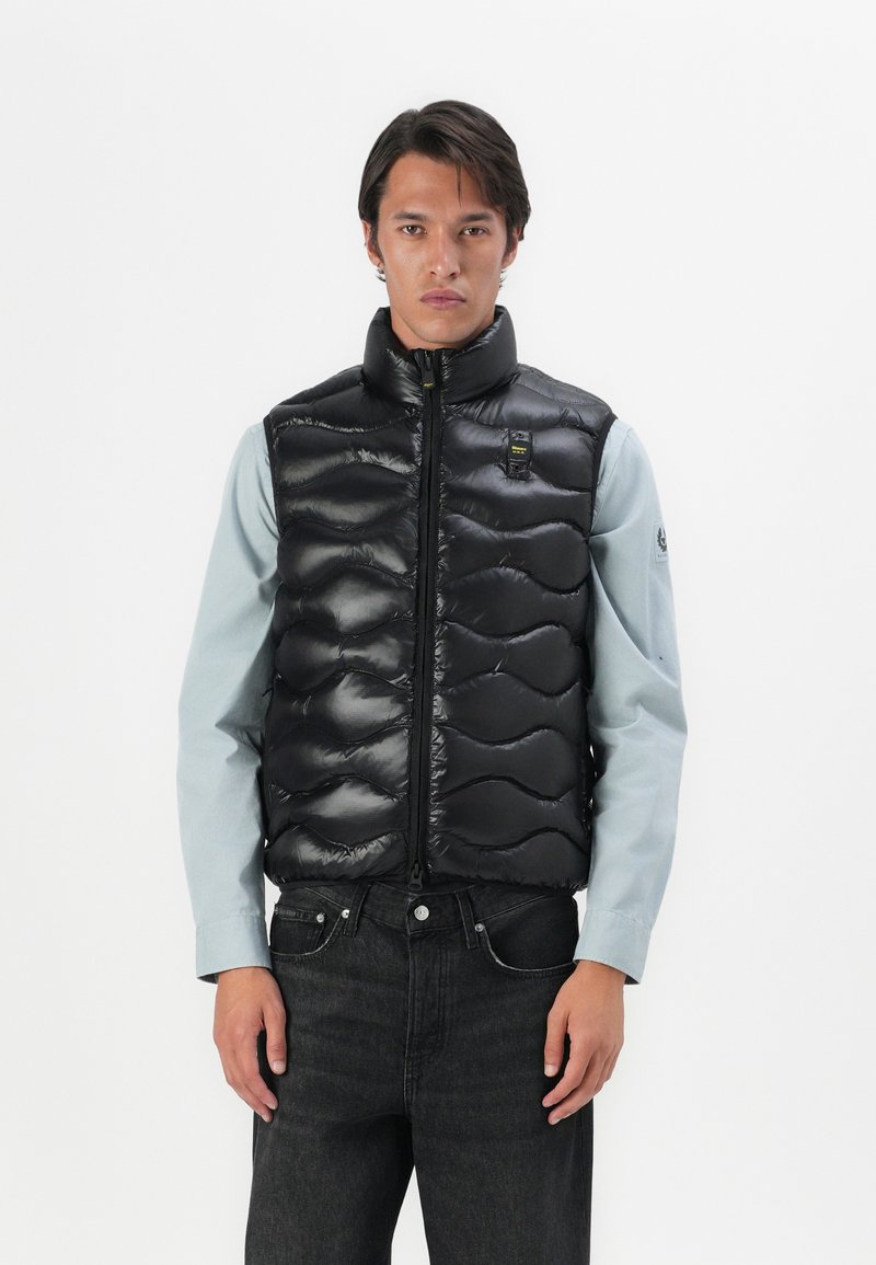 Black puffer vest with a high neck, quilted texture, and front zipper. Worn over a light blue shirt, paired with dark jeans.