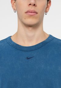 Plava pamučna majica s okruglim izrezom, sa malim tamnoplavim Nike logotipom na prsima. Tekstura izgleda blago izblijedjela.