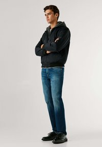 Chaqueta bomber negra con cremallera y capucha gris, usada sobre una camisa blanca. Vaqueros azules y zapatos negros con cordones. Posición de pie con los brazos cruzados.