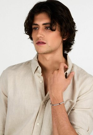 Giovane uomo con capelli scuri che indossa una camicia beige con bottoni, toccandosi il colletto e indossando un braccialetto d'argento con perle tonde al polso sinistro.