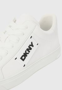 Bijeli tenisice s glatkim sintetičkim gornjim dijelom, okruglim vrhom, ravnim vezicama i crnim DKNY logotipom na strani. Gumeni đon.