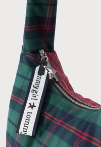 Sac à épaule en tissu plaid de couleur verte, rouge et marine. Il dispose d'une fermeture éclair argentée, d'une tirette en cuir noir et d'une étiquette décorative rayée avec du texte.