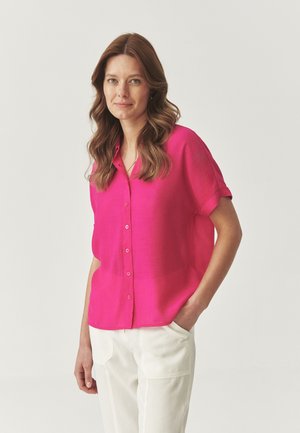 Camicia fucsia con bottoni, maniche corte e vestibilità rilassata, abbinata a pantaloni bianchi. Tessuto morbido, tessuto leggero e design semplice.
