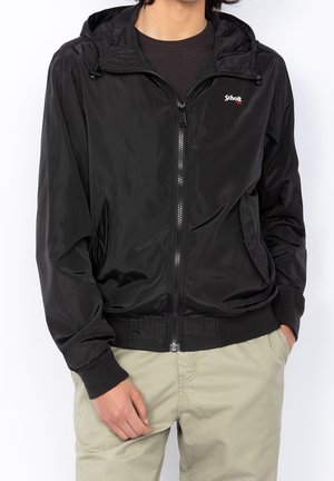 Homme portant une veste noire zippée avec capuche et un pantalon beige, les mains dans les poches, debout devant un fond blanc uni.