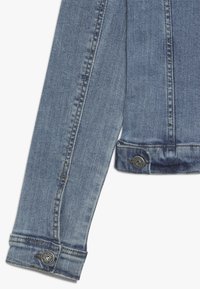 Džínová bunda ve světle modré barvě, s klasickým střihem, knoflíkovými detaily na manžetách a viditelným šitím podél švů. Měkká textura.