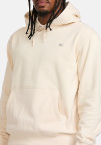 Cremefarbener Hoodie aus weichem Material, mit einer Fronttasche, Kapuze mit Kordelzug und einem kleinen Logo-Patch auf der Brust.
