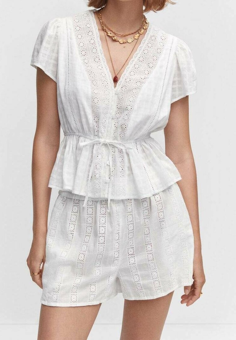 Femme portant un chemisier blanc à manches courtes avec col en V, ceinture nouée à la taille et short en dentelle taille haute assorti, accessoirisé de colliers dorés et rouges superposés.