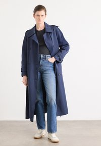 Mørkeblå trenchcoat med skulderepauletter, kombinert med høytlivs blåjeans og hvite joggesko med gummisåler. Minimalistisk design, glatt tekstur.