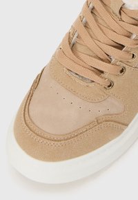 Baskets en suède tan avec des accents beige, présentant un bout arrondi, des détails perforés et une semelle en caoutchouc blanche. Les lacets sont marron clair avec des œillets.