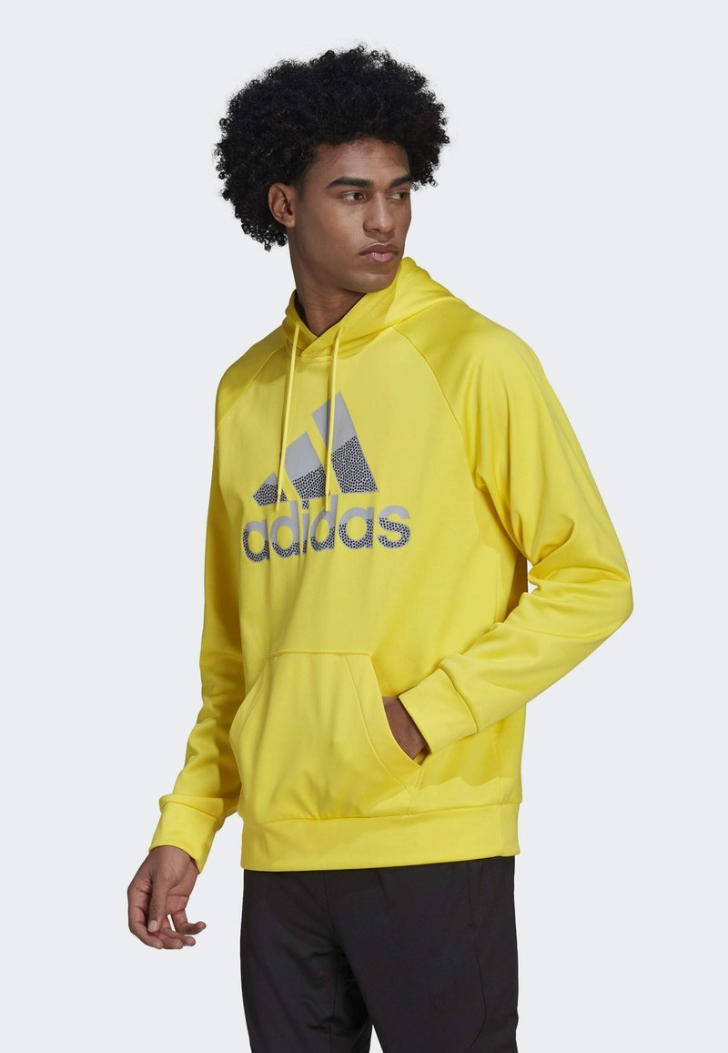 Felpa gialla con cappuccio e lacci, tasca frontale e logo Adidas grigio. Presenta una texture liscia e un design aderente.