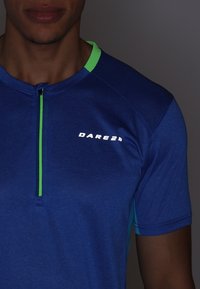 Camisa atlética de manga corta azul con una cremallera en la parte delantera, acentos verdes en el cuello y un logo reflectante en el pecho.