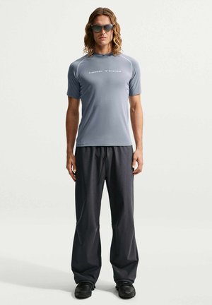 Homme aux longs cheveux portant des lunettes de soleil foncées, un t-shirt gris Nike Tech, un pantalon noir ample et des chaussures noires, debout devant un fond clair uni.