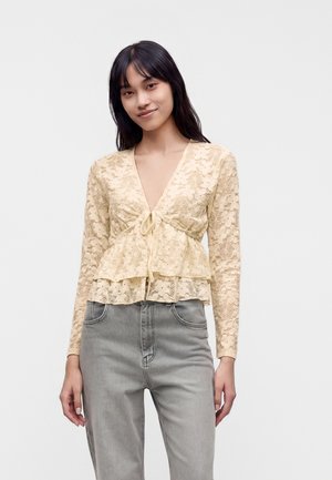 Jeune femme portant un haut à manches longues en dentelle florale beige avec un nœud devant et un jean taille haute gris clair, debout devant un fond uni.
