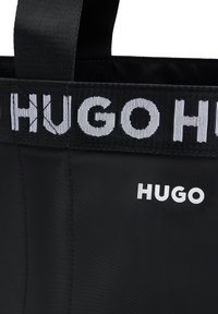 Borsa tote nera realizzata in materiale sintetico liscio, con un audace logo bianco "HUGO" sul davanti e una tracolla intrecciata nera a contrasto.