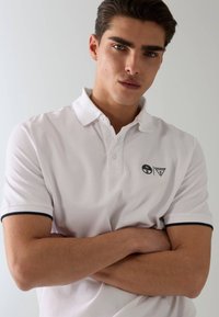 Jeune homme les bras croisés, portant un polo blanc avec des manches bordées de noir et un petit logo sur la poitrine, debout devant un fond uni.