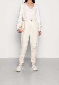 Pull col en V crème à manches longues, associé à un pantalon crème fuselé et taille haute. Baskets blanches avec des accents gris et un sac beige.