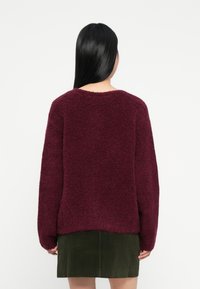 Mulher com cabelo preto longo, vestindo um suéter felpudo bordeaux e uma saia de veludo cotelê verde escuro, de costas, em um fundo branco simples.