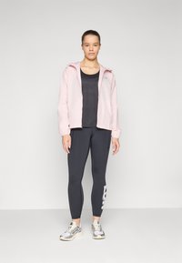 Jachetă cu fermoar de culoare roz pal, cu glugă, top sportiv negru, colanți închiși la culoare cu modele pe lateral și adidași gri cu accente galbene.