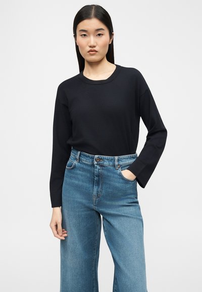 Femme aux cheveux noirs raides portant une chemise noire à manches longues et un jean bleu taille haute, une main dans la poche.