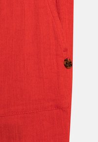 Czerwona teksturowana tkanina z widocznymi szwami i małym okrągłym brązowym guzikiem przyszytym w pobliżu krawędzi.