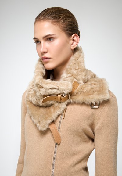 Giovane donna con capelli tirati indietro, che indossa un maglione beige con zip e un ampio colletto in pelliccia sintetica beige con fibbie in pelle.