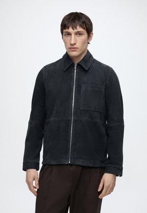Joven con una chaqueta de ante negra con cremallera y bolsillo en el pecho y pantalones marrón oscuro, de pie frente a un fondo claro y liso.