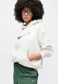Sweatshirt branco com capuz e logótipo azul da Nike, com um bolso canguru e punhos canelados. Usado com calções desportivos verdes.