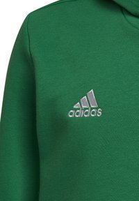 Zielony materiał z białym haftowanym logo Adidasa, które przedstawia trzy ukośne paski oraz nazwę marki.