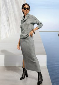 Frau in grauem Strickpullover und Rock mit Schlitz, schwarzen Stiefeletten, Sonnenbrille und geometrischem Armband, die am Wasserstand steht.