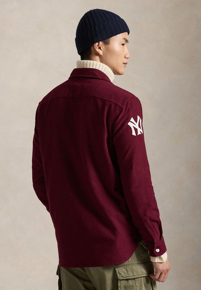 Μπορντό πουκάμισο με κουμπιά και λευκό λογότυπο NY στην αριστερή μανσέτα, συνδυασμένο με ναυτικό μπλε ριγωτό σκουφάκι και κρεμ χούφτα.